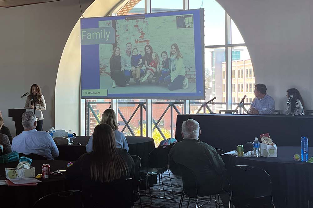 A woman stands at a podium on the far left in front of a screen with a photo of a family. People in the audience are watching her.